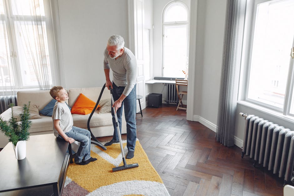 A yellow canister vacuum cleaner with a flexible hose and cleaning head is positioned on a decorative rug in a living room. The room features wooden flooring and a low black metal sideboard with a woo