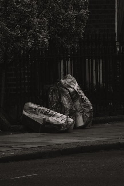 Photograph depicts two large black plastic rubbish bags filled with waste, positioned on a concrete sidewalk beside a black metal fence. The bags appear to contain bulky waste items, possibly packagin