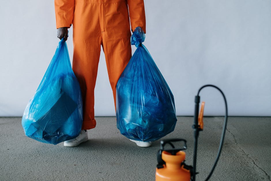 Photograph depicts two large black plastic rubbish bags filled with waste, positioned on a concrete sidewalk beside a black metal fence. The bags appear to contain bulky waste items, possibly packagin