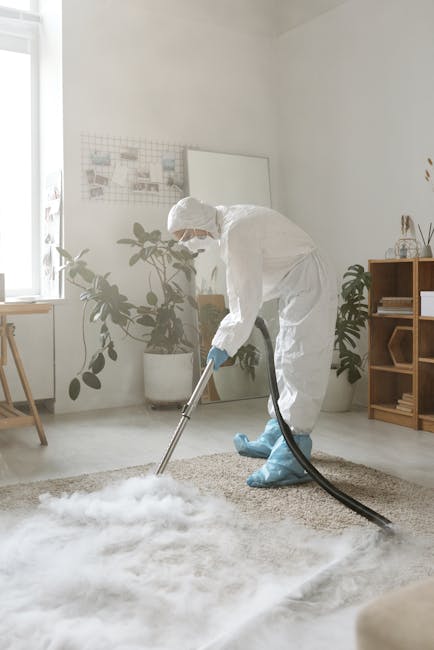 A professional cleaning specialist dressed in white protective coveralls, a face mask, and blue gloves, performing steam or deep cleaning on a beige carpet in a modern living room. The room features a