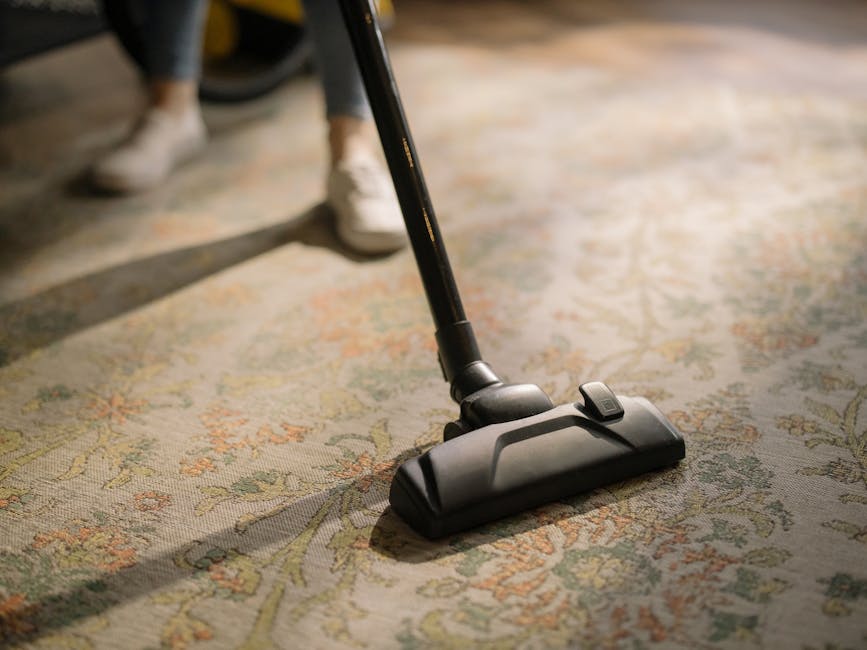 A woman with curly hair, dressed in a beige jacket and blue jeans, is engaged in surface cleaning using a vacuum cleaner on a traditional patterned area rug in a living room. The room features wooden